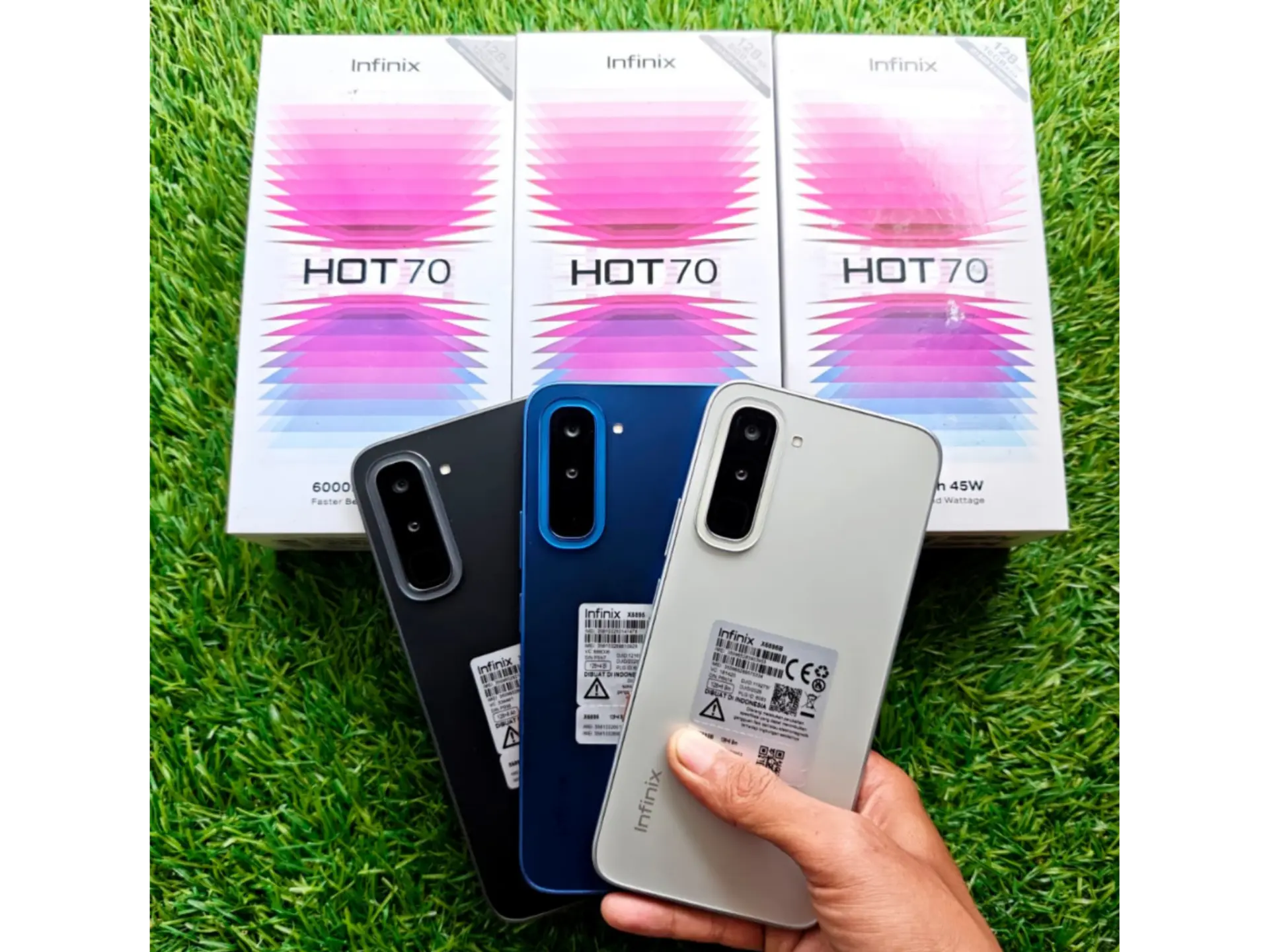 Three Infinix Hot 70 phones in black, blue and white held over grass in front of their retail boxes — 128GB, 6000mAh, 45W charging